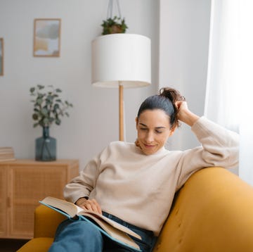 view of a cheerful woman sitting on a sofa in her living room reading a book. concept of rest and leisure activity
