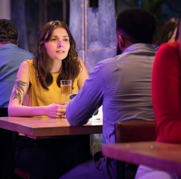 a woman in a yellow shirt looks concerned in the middle of speaking to her date. he sits with his back to the camera. they are in the middle of a crowded and stylish restaurant, bathed in purple light. behind them two people look at a phone. the cocktail in front of her is served in a modern glass, and she has a tattoo.