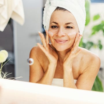 a smiling woman with a towel wrapped around her head applies facial cream in front of a bathroom mirror. natural light and green plants convey a fresh, at home skincare moment.